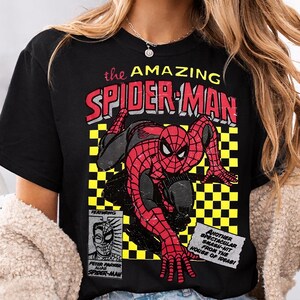 May include: Black t-shirt with a comic book style graphic of Spider-Man in red and black. The text "the AMAZING SPIDER-MAN" is above the image. Additional text includes "FEATURING PETER PARKER ALIAS SPIDER-MAN" and "ANOTHER SPECTACULAR SMASH-HIT FROM THE HOUSE OF IDEAS!"