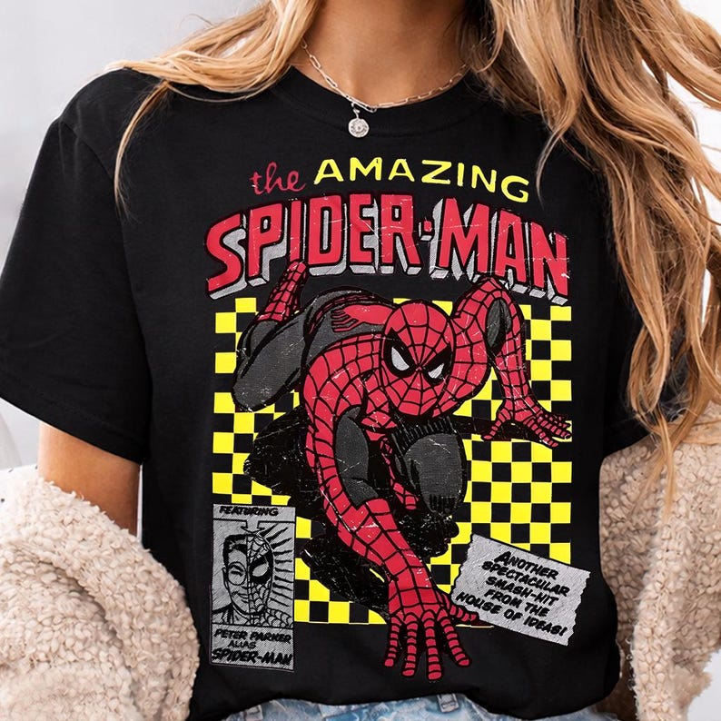 May include: Black t-shirt with a comic book style graphic of Spider-Man in red and black. The text "the AMAZING SPIDER-MAN" is above the image. Additional text includes "FEATURING PETER PARKER ALIAS SPIDER-MAN" and "ANOTHER SPECTACULAR SMASH-HIT FROM THE HOUSE OF IDEAS!"