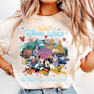 K&ouml;nnte beinhalten: Cremefarbenes T-Shirt mit einer farbenfrohen Grafik von Disney-Figuren und Wahrzeichen. Der Text "Walt Disney World" ist oben gew&ouml;lbt, darunter "The Most Magical Place on Earth". Micky, Minnie, Donald, Daisy und Goofy sind abgebildet.