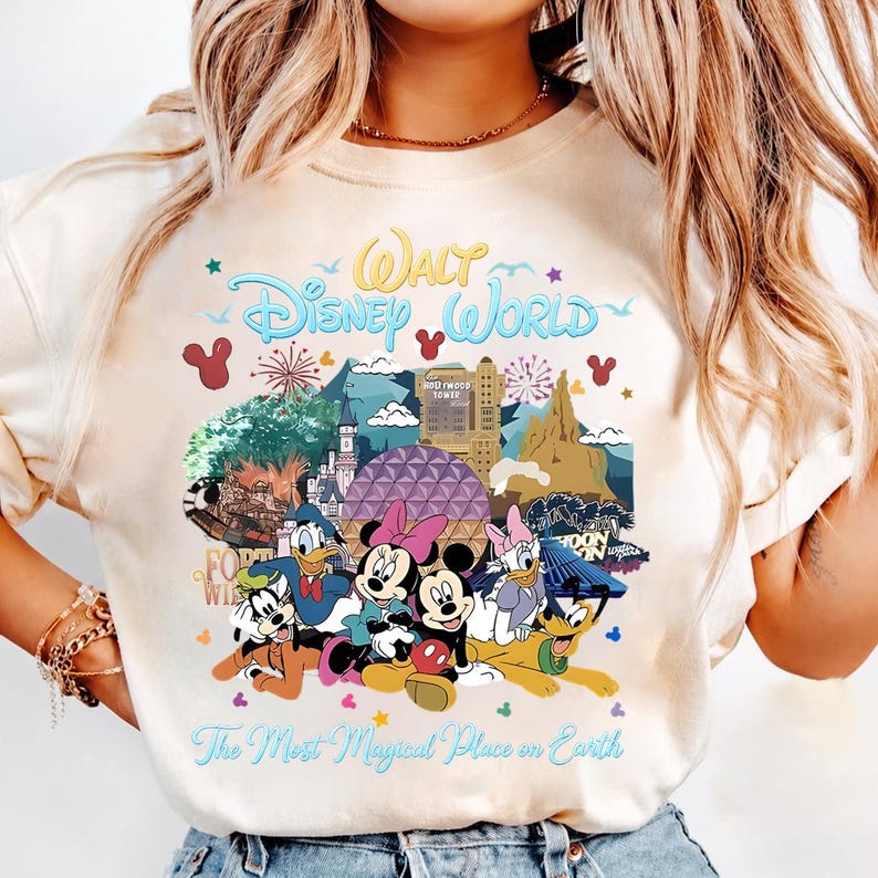 K&ouml;nnte beinhalten: Cremefarbenes T-Shirt mit einer farbenfrohen Grafik von Disney-Figuren und Wahrzeichen. Der Text "Walt Disney World" ist oben gew&ouml;lbt, darunter "The Most Magical Place on Earth". Micky, Minnie, Donald, Daisy und Goofy sind abgebildet.