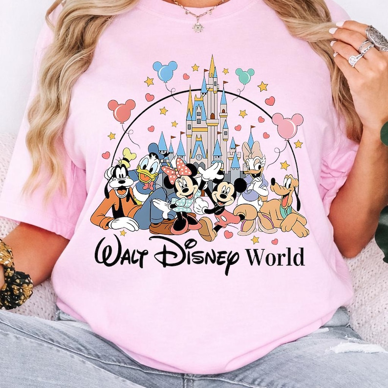 K&ouml;nnte beinhalten: Hellrosa T-Shirt mit einer Grafik von Disney-Figuren, darunter Mickey und Minnie Mouse, Goofy, Donald und Daisy Duck und Pluto. Die Worte "Walt Disney World" sind unter den Figuren aufgedruckt.