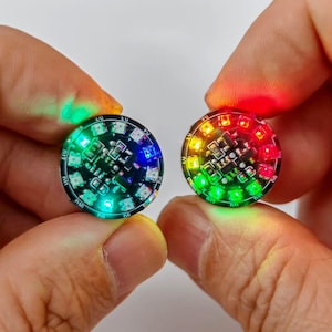 Puede incluir: Dos placas de circuito electr&oacute;nico circulares con luces LED integradas. La placa izquierda emite un brillo verde y azul intenso, mientras que la derecha muestra un patr&oacute;n de luz rojo, naranja y verde sobre sus componentes.