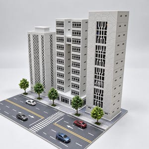 Peut inclure: Une maquette de trois immeubles blancs de grande hauteur avec des motifs de façade géométriques distincts, placés à côté d'une route en asphalte gris avec des marquages blancs, un passage piéton et quatre voitures miniatures.