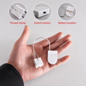 May include: A white plastic battery-operated light component featuring a small rectangular thread clamp, a switch button, and a battery compartment. The device is connected by thin clear wires, with circular insets showing the thread clamp, switch button, and battery location.