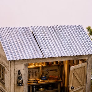 Peut inclure: Un abri miniature en bois avec un toit en métal ondulé, révélant un établi avec des outils, une lanterne et un baril vert. La structure comprend une porte en bois, un chemin en pierre, un tas de bois et une petite clôture.