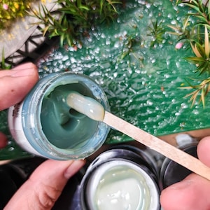 May include: A jar of teal-colored paint with a wooden stir stick. The paint is thick and creamy, and the jar is partially open. Another jar of paint is visible in the foreground.