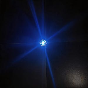 Può includere: Una piccola sorgente luminosa circolare che emette un bagliore blu brillante e intenso. La luce si irradia verso l'esterno in quattro fasci distinti e nitidi che formano un motivo a croce su uno sfondo scuro e uniforme. Il centro è strutturato.