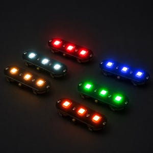 May include: Five small, elongated, black electronic modules, each featuring three integrated LED lights. The modules are arranged in a cluster, with each set emitting a distinct colour: red, white, blue, green, and orange, all glowing against a dark background.