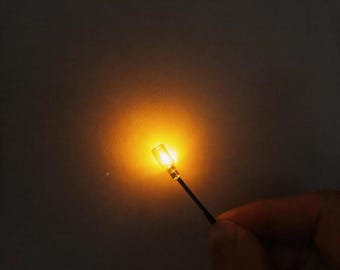Puede incluir: Un pequeño componente de luz LED cilíndrico que emite un brillo naranja cálido y brillante. La luz está unida a un cable negro delgado, creando un punto de iluminación enfocado contra un fondo oscuro y neutro, resaltando el diodo.