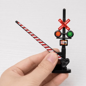 May include: A miniature black plastic railroad crossing signal with a red and white striped barrier arm. The signal features a red crossbuck sign with the text RAILROAD, two circular lights in red and green, and a small square sign with a train icon.
