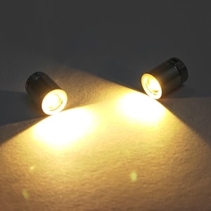 May include: Two small, cylindrical metal spotlights emitting a warm, bright yellow light. Each fixture features a circular lens at the front, casting a focused beam of illumination outward, creating a distinct glow on the surface below.