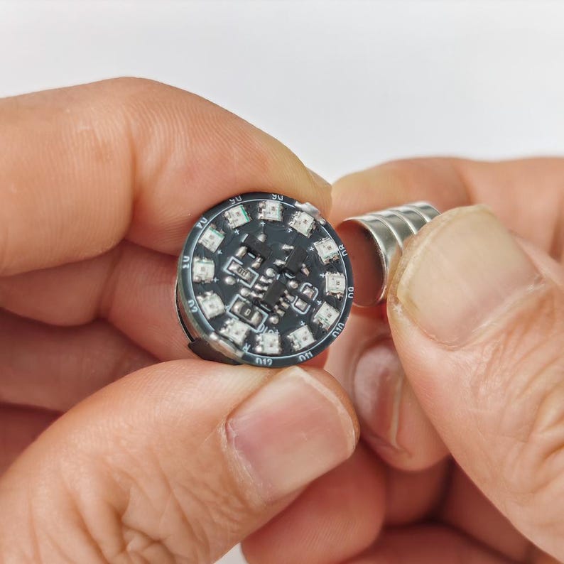 Puede incluir: Una placa de circuito impreso circular negra con m&uacute;ltiples componentes LED peque&ntilde;os montados en superficie dispuestos alrededor del per&iacute;metro, con chips electr&oacute;nicos integrados y puntos de contacto met&aacute;licos plateados visibles en la superficie.