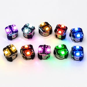 Peut inclure: Un ensemble de dix petits modules lumineux LED de forme carrée disposés sur deux rangées. Chaque module présente une base de circuit imprimé noire avec deux diodes électroluminescentes intégrées qui brillent en violet, blanc, jaune, rouge, bleu et vert.