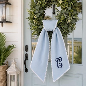 May include: A light blue fabric door hanger with a white scalloped edge and a dark blue embroidered letter C monogram. The hanger is draped over a green foliage wreath hanging on a light blue door with a black handle and a lantern.
