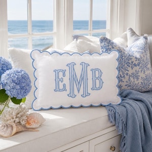 May include: A rectangular white throw pillow with a scalloped edge trimmed in light blue. The center features a large, embroidered monogram with the letters E, M, and B in a light blue striped pattern. The pillow is made of a smooth fabric.