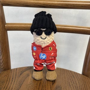 May include: A plush doll with a cream-colored textured body wearing a black knit beanie, black round sunglasses, and a red racing jumpsuit featuring various sponsor logos including HP and the Ferrari emblem, with tan corduroy-textured feet.