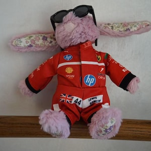 May include: A plush pink rabbit toy wearing black sunglasses and a red racing jumpsuit. The jumpsuit features various sponsor logos, the number 44, a British flag, and the name Lewis Hamilton printed on the front. The rabbit has floral fabric ears.