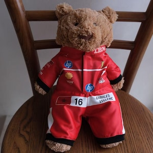 May include: A brown plush teddy bear wearing a red racing jumpsuit with white accents. The suit features logos including IBM, HP, and Shell, along with the number 16 and the name Charles Leclerc printed on the front in black and white text.