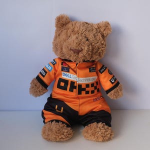 Könnte beinhalten: Ein brauner Plüsch-Teddybär in einem leuchtend orange-schwarzen Rennanzug. Der Anzug ist mit verschiedenen Sponsorenlogos wie OKX, Dell Technologies und Cisco bedruckt, hat schwarze Ärmel und Hosenbeine sowie eine kleine britische Flagge.