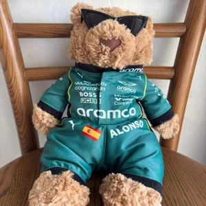 May include: A tan plush teddy bear wearing black sunglasses and a green racing jumpsuit. The jumpsuit features the text Aramco, Alonso, Boss, JCB, and Cognizant, along with a small Spanish flag patch on the chest and a white Puma logo on the side.