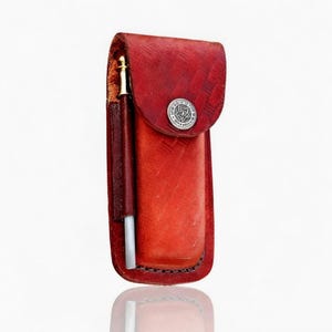 Puede incluir: Un estuche de cuero rojo para bol&iacute;grafo con un emblema plateado en la solapa. Se ve un bol&iacute;grafo con punta dorada en el bolsillo lateral. El estuche tiene un borde cosido y un acabado texturizado.