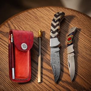 May include: A collection of knives and accessories on a wooden surface. Includes a red leather case with a pen, two knives with patterned blades and decorative handles, and a sharpening rod with a gold-colored top.