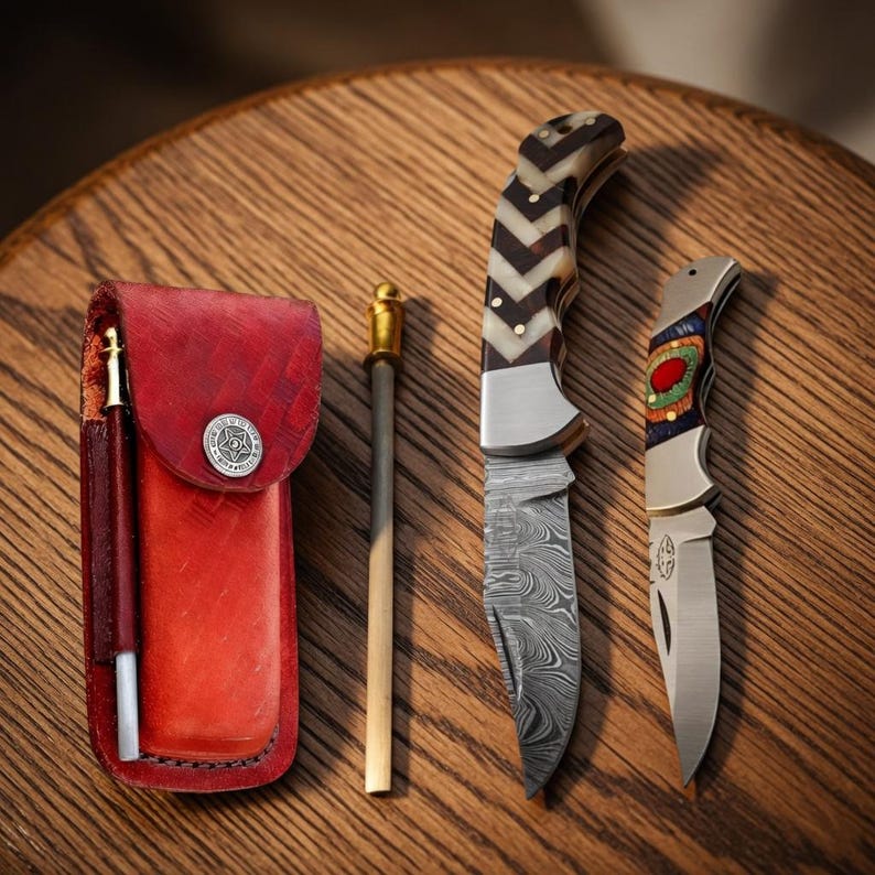 Handmade knife set on dark wooden table with brown leather sheath, sharpening rod, Damascus-style fixed blade knife, and folding pocket knife with decorative handle.