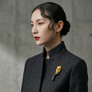 May include: A gold-colored mask brooch with a black base is pinned to a dark gray jacket. The brooch is a stylized representation of a face. The jacket has a high collar and is worn by a person with dark hair and red lipstick.