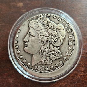 Peut inclure: Une pièce de monnaie Morgan dollar argentée datée de 1884, enfermée dans une capsule de protection circulaire en plastique transparent. L'avers présente le profil de Lady Liberty tourné vers la gauche, avec le texte LIBERTY, E PLURIBUS UNUM.
