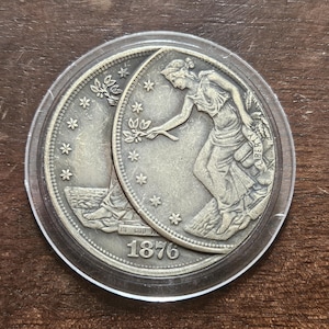 Könnte beinhalten: Eine silberfarbene Münze mit der Jahreszahl 1876, eingeschlossen in einer transparenten, runden Schutzhülle. Die Münze zeigt das Relief einer stehenden Figur, die einen Zweig hält, umgeben von Sternen, mit dem Wort LIBERTY auf einem Banner.