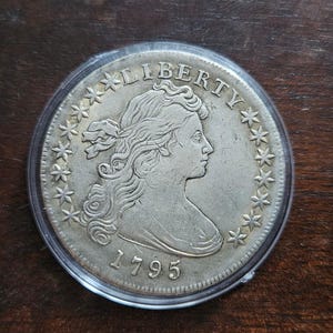 Peut inclure: Une pièce de monnaie argentée enfermée dans un étui de protection en plastique transparent. L'avers présente un portrait de profil tourné vers la droite, avec le mot LIBERTY inscrit au-dessus et l'année 1795 en dessous, entouré de quinze étoiles.