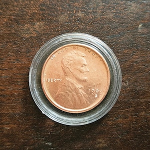 Peut inclure: Une pièce de monnaie américaine d'un cent, en cuivre, représentant le profil d'Abraham Lincoln. La pièce est datée de 1999, porte les inscriptions IN GOD WE TRUST et LIBERTY, et est insérée dans un étui de protection circulaire en plastique.