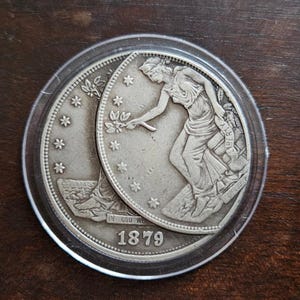 Puede incluir: Una moneda de tono plateado que muestra una figura sentada sosteniendo una rama y una pancarta con la palabra LIBERTY, con el año 1879 visible en la parte inferior. La moneda está dentro de un estuche protector circular de plástico.