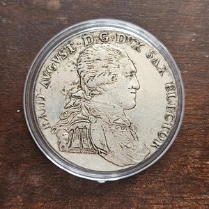 May include: A silver-toned circular coin encased in a clear protective plastic capsule. The coin features a profile portrait of a person in 18th-century attire with the text FRID AVGUST D G DUX SAX ELECTOR embossed along the outer circular edge.