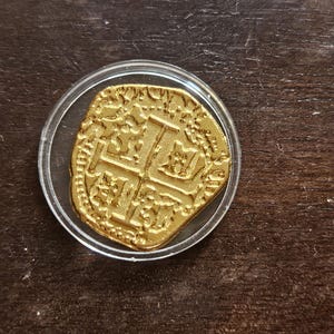 Puede incluir: Una moneda de color dorado con una forma irregular y redondeada, con un diseño de cruz en relieve en su superficie, encerrada dentro de una cápsula protectora de plástico transparente y circular. La moneda presenta detalles en relieve y un acabado metálico.