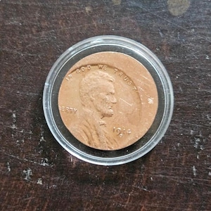 Pode incluir: Uma moeda de um centavo Lincoln de 1914, cor de cobre, dentro de um estojo protetor circular de plástico transparente. A moeda apresenta o perfil de Abraham Lincoln voltado para a direita, com o texto IN GOD WE TRUST e o ano 1914 visíveis.