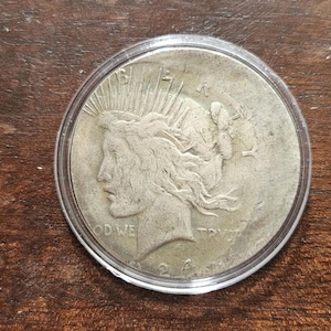 May include: A silver Peace dollar coin encased in a clear plastic protective capsule. The obverse side features the profile of Lady Liberty wearing a radiant crown, with the text LIBERTY, IN GOD WE TRUST, and the year 1924 visible on the coin surface.