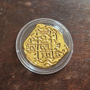 Puede incluir: Una moneda de color dorado con una forma irregular y no circular, encerrada en una cápsula protectora de plástico redonda y transparente. La moneda presenta marcas en relieve, incluido el número 2 y varios símbolos y líneas estilizadas.