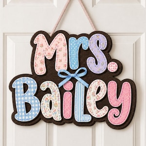 May include: A dark brown wooden wall sign featuring the text Mrs. Bailey in layered, multi-colored fabric-covered letters. The letters display pink floral, blue polka dot, purple floral, blue gingham, and pink patterns with a light blue ribbon bow.