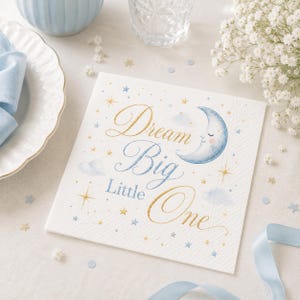May include: A square white paper napkin featuring the text Dream Big Little One in gold and blue script. The design includes a blue crescent moon with a face, light blue clouds, and scattered gold and blue stars on a textured white background.