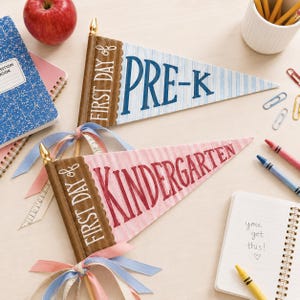 Op de afbeelding: Twee driehoekige viltwimpels met houten stokjes en gouden knoppen. De ene wimpel is lichtblauw met witte strepen en de tekst "FIRST DAY OF PRE-K" in donkerblauw. De andere is lichtroze met witte strepen en de tekst "FIRST DAY OF KINDERGARTEN" in donkerrood, beide versierd met pastelkleurige linten.