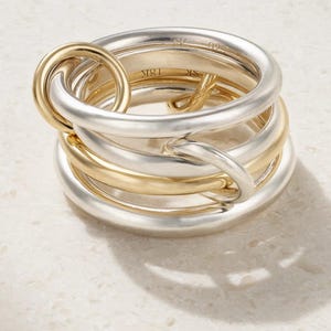 Peut inclure: Une bague multi-bandes combinant argent et or. La bague présente un design unique avec des bandes entrelacées et une boucle dorée proéminente. Les bandes sont lisses et polies, créant un look moderne et élégant. Un bijou sophistiqué.