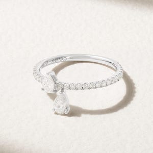 May include: A silver-toned ring featuring a thin band encrusted with small round clear stones. The ring has an open design with two pear-shaped clear stones dangling from the band. The interior of the band is engraved with the text ANITA KO.