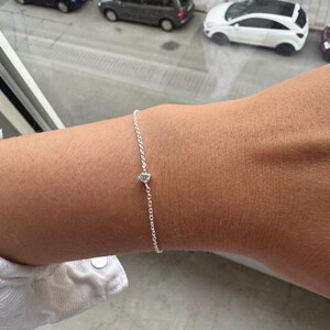 Pode incluir: Uma delicada pulseira de corrente de prata com uma única pedra preciosa pequena, redonda e transparente, fixada num suporte de metal simples com garras. A pulseira é fina e tem um acabamento metálico subtil e refletor sobre a pele.