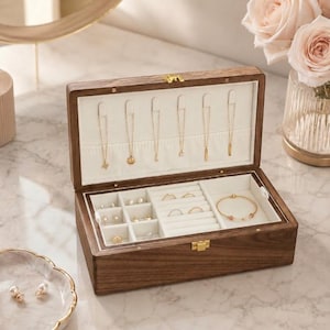 May include: Open wooden jewelry box with a light-colored interior and multiple compartments. The box is filled with gold necklaces, rings, earrings, and a bracelet. The box has a gold clasp and hinges.