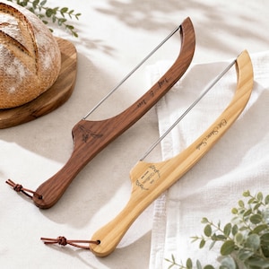 Op de afbeelding: Twee houten broodsnijders met roestvrijstalen messen. Eén is donkerbruin met de tekst "Leaf & Love", de andere is lichtbruin met de tekst "Eat, Share, Smile". Beide hebben leren bandjes.