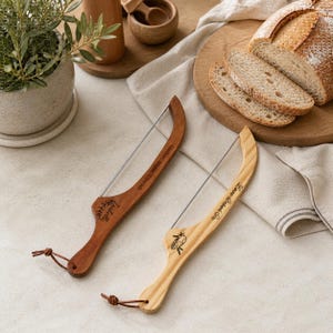 May include: Two wooden bread slicers with wire blades. One is dark brown with the name "Isabella" and the text "Gather, Share, Cherish". The other is light brown with the text "Love Baked On" and a small graphic.
