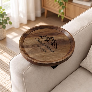 May include: A round wooden sofa arm tray with a dark brown finish and a raised edge. The surface features a laser-engraved mountain graphic above the text Trisha & Michael and the date 10/2/2026, designed to rest securely over a couch armrest.