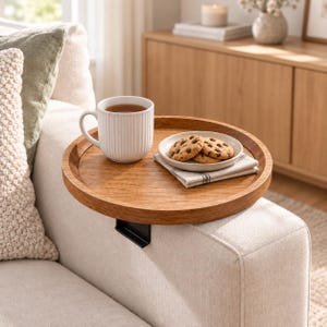 May include: A round wooden armrest tray with a black metal clamp attachment. The tray holds a white ribbed ceramic mug filled with a brown beverage and a small white plate containing three chocolate chip cookies on a folded fabric napkin.