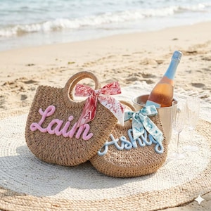 May include: Two woven straw tote bags with handles, personalized with the names "Lain" in pink and "Ashly" in blue. Each bag has a decorative ribbon. A bottle of ros&eacute; in an ice bucket and two champagne flutes sit on a beach blanket.